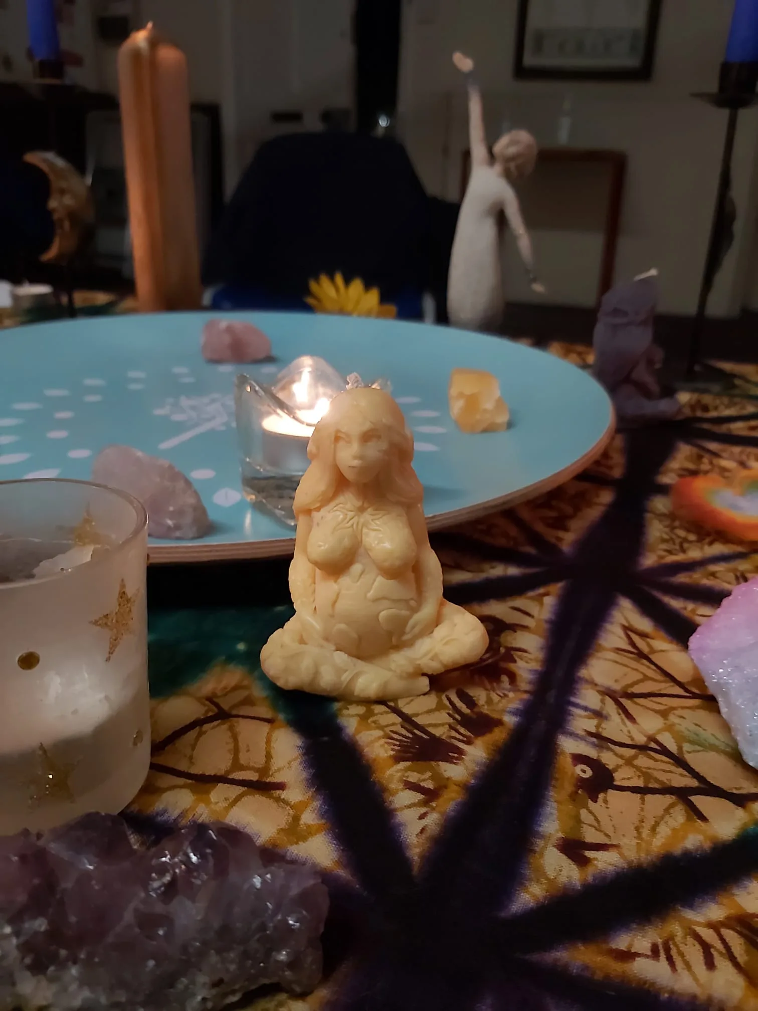 The image depicts a decorative setup on a table with various items. In the center, there is a small candle shaped like a figure of a seated woman. Around it, there are several crystals and stones placed on a patterned cloth. The background includes a tall candle and abstract figurines. A serene and mystical ambiance is suggested by the arrangement and lighting.